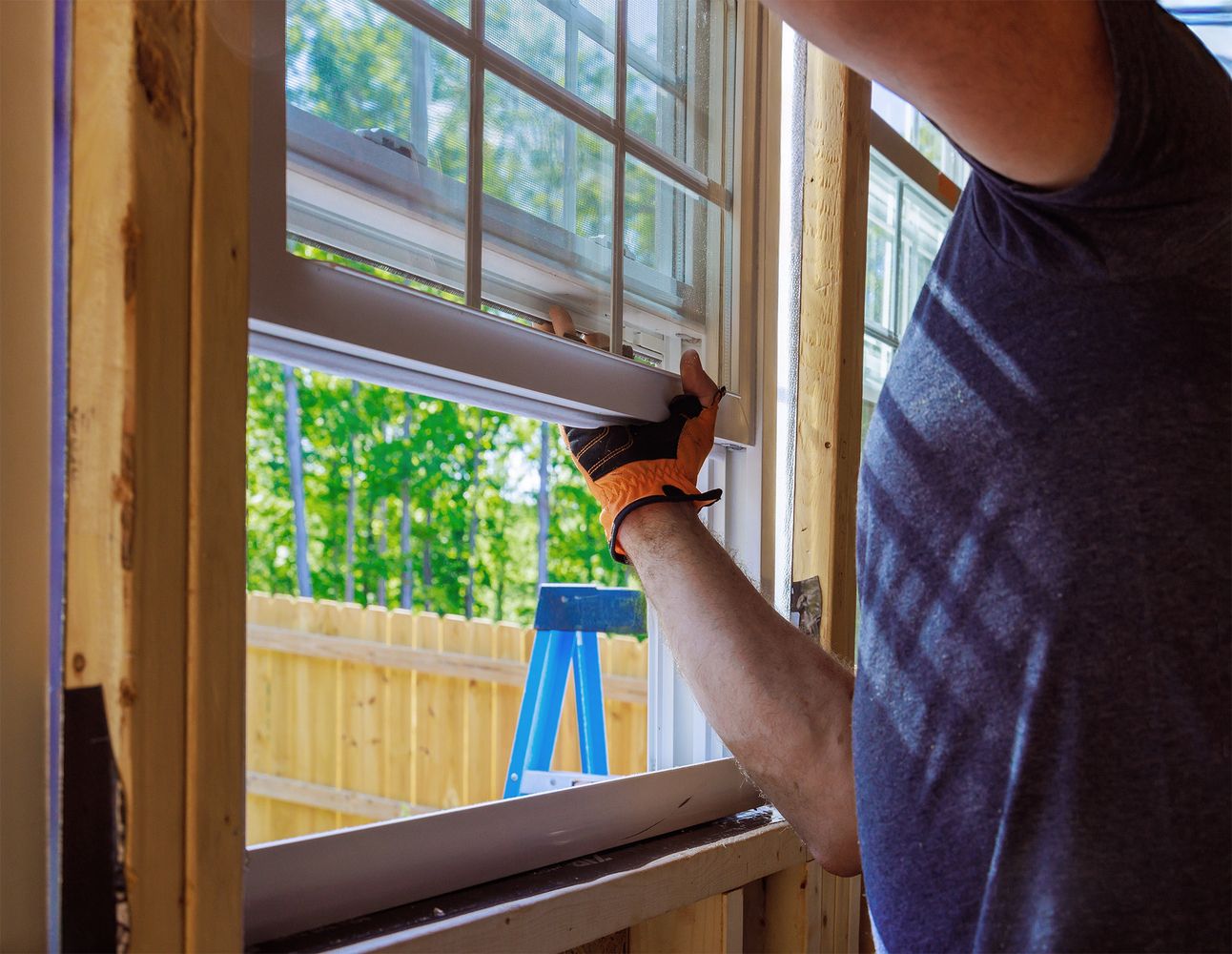 Eine Person setzt ein Fenster in einen offenen, gelben Holzrahmen, draußen sind Bäume und eine blaue Leiter sichtbar.